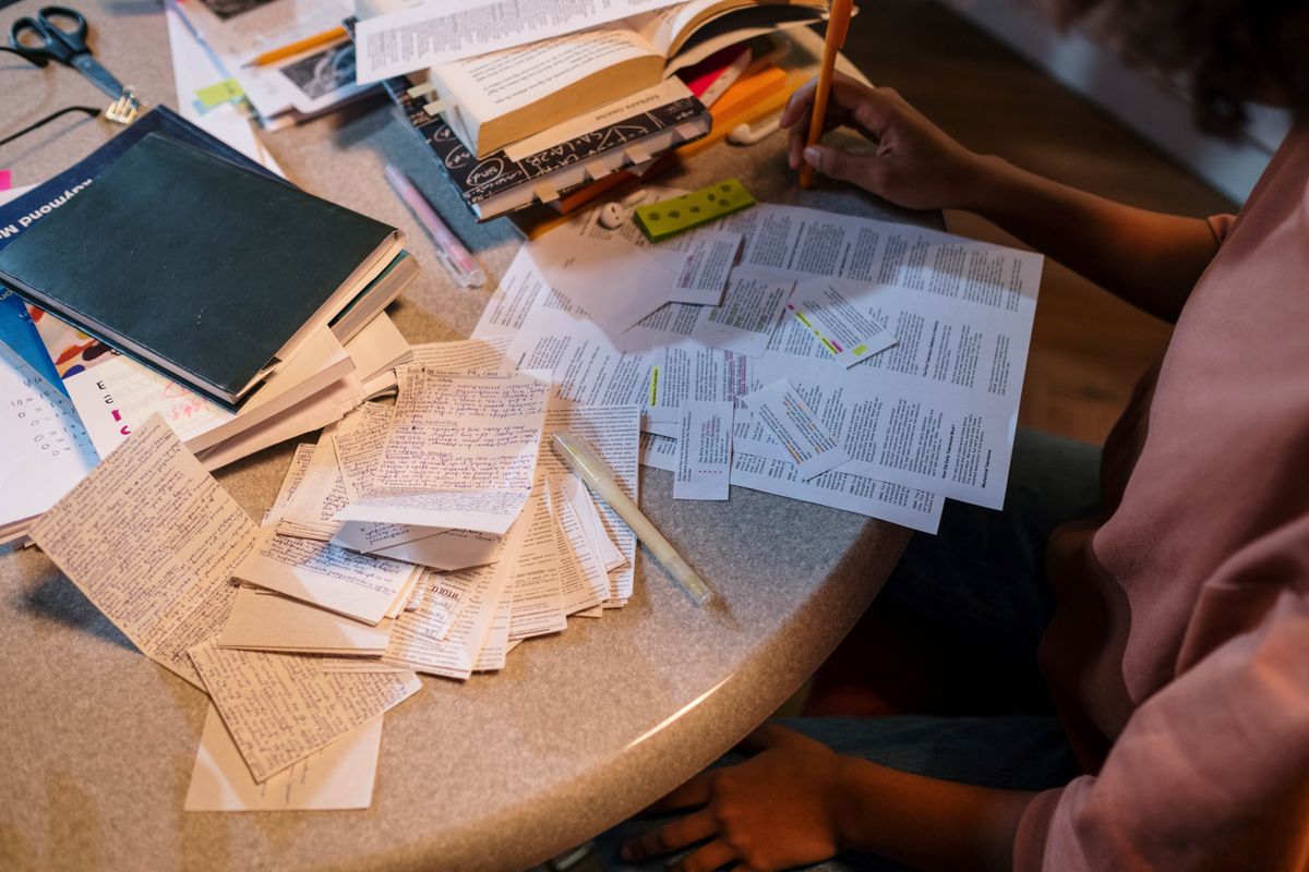 A round table seen from above, spread with dozens of small handwritten revision cards, printed pages cut into strips, stacked books tabbed with folded paper, and a hand at the edge of the frame holding a pencil