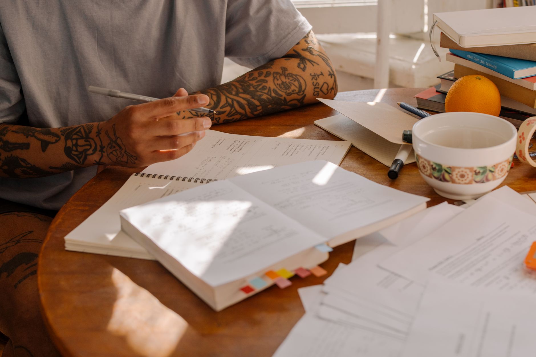 A person in a gray t-shirt with tattooed forearms sits at a sunlit wooden table, pen in hand, over a spiral pad of handwritten notes and a thick book flagged with coloured tabs, with a stack of textbooks and a bowl at the far edge