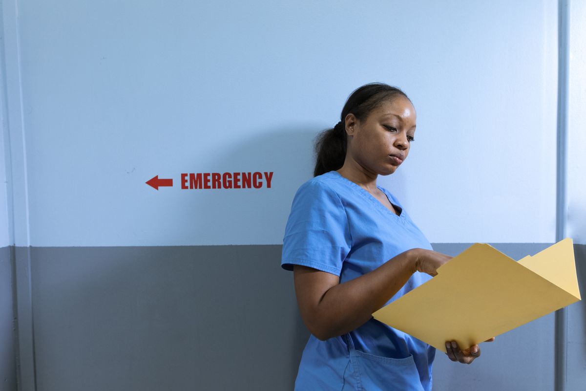 A nurse in blue scrubs leaning against a corridor wall reading an open patient folder, beneath a hospital sign for the emergency department