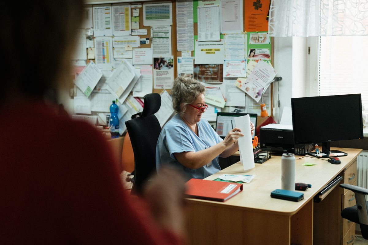 A nurse in blue scrubs sitting at a hospital office desk, sorting a stack of printed records into piles beside a keyboard and a mug