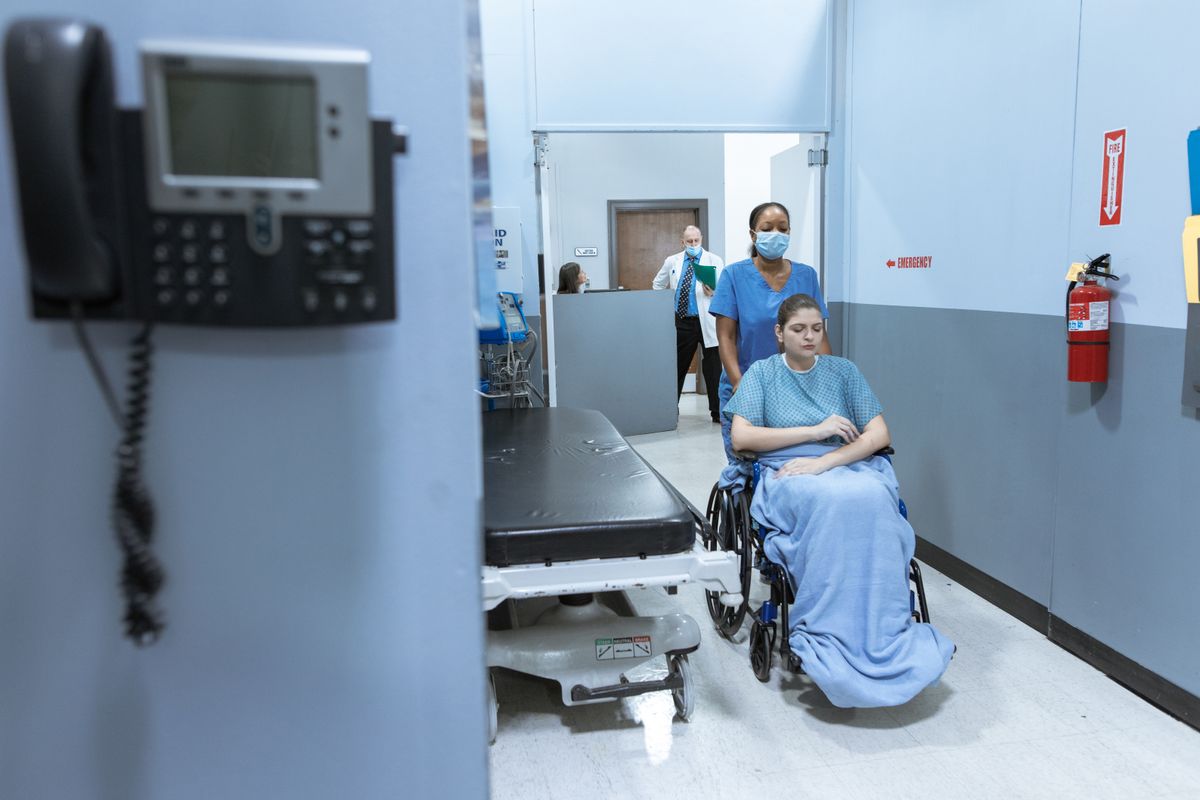 A masked nurse in blue scrubs pushes a patient wrapped in a blanket in a wheelchair along a hospital corridor, past a parked gurney, while a clinician holding a chart stands further down the hall beneath a sign pointing to the emergency department