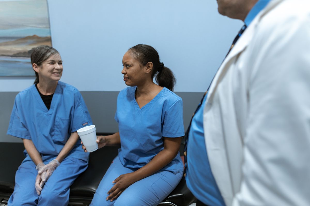 Two nurses in scrubs talking during a break in a hospital corridor, one holding a drink