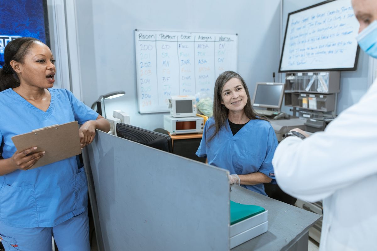 Two nurses in blue scrubs at a hospital counter, one holding a clipboard and speaking, with a whiteboard behind them ruled into columns headed room, patient, code, allergies and nurse, and a masked colleague in a white coat at the edge of the frame