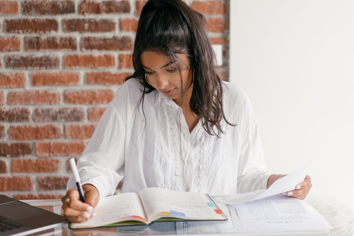 A woman in a white shirt writes in a tabbed notebook at a glass-topped table against a brick wall, holding a printed page up in her other hand, a closed laptop at her elbow